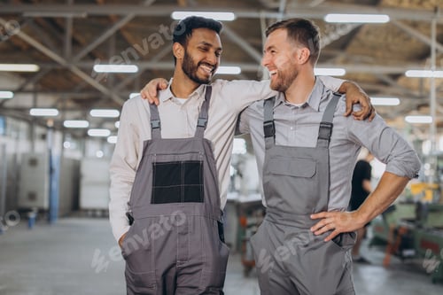 Preview: Two happy international workers in overalls hug each other against a factory background