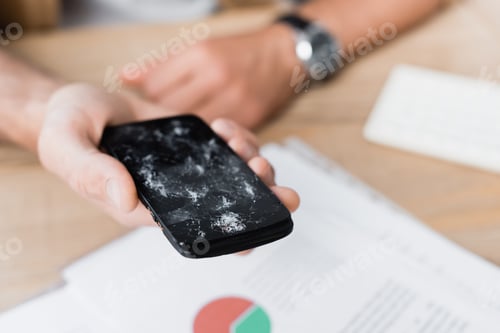 Preview: Cropped view of businessman holding smashed smartphone with blurred workplace on background