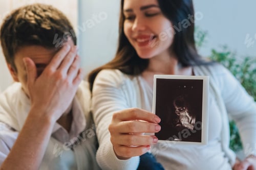 Preview: Young couple waiting for baby. Happy pregnant woman showing pregnancy ultrasound to husband