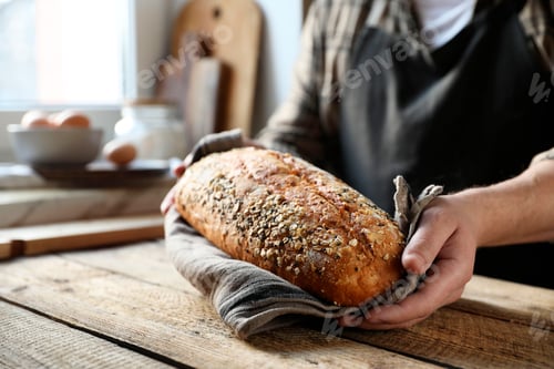 Preview: Man holding loaf of fresh bread at wooden table indoors, closeup