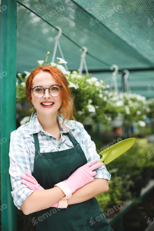 Preview: Beautiful florist in apron and pink gloves standing with little garden shovel in hand