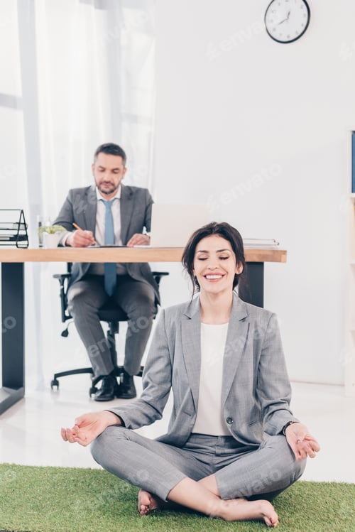 Preview: smiling businesswoman meditating in Lotus Pose on grass mat while businessman sitting at table and