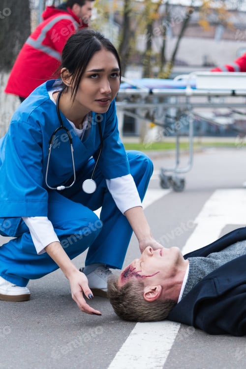 Preview: young female Asian doctor helping unconscious injured man lying on a street