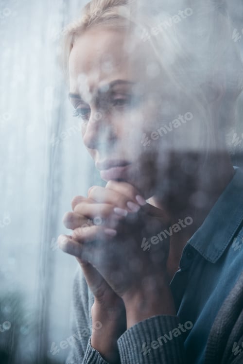 Preview: sad adult woman with folded hands at home through window with raindrops