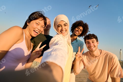 Preview: Happy multi ethnic friends taking selfie, showing peace sign at sunset