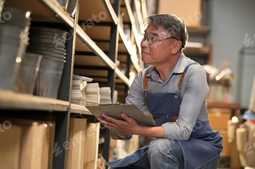 Preview: Elderly Asian man in apron checking inventory on shelves with clipboard