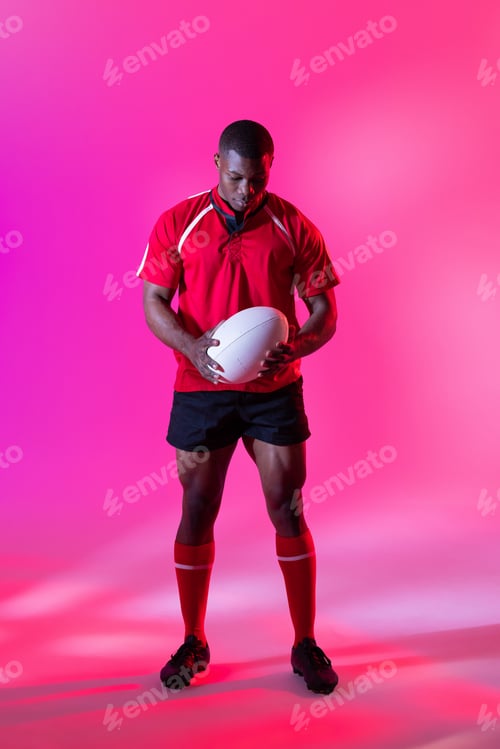 Preview: African american male rugby player with rugby ball over pink lighting