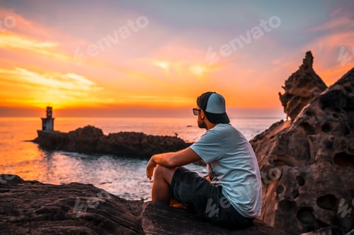 Preview: A young man sitting in a white shirt and cap looking at the orange sunset
