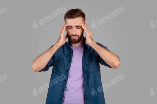 Preview: Young man suffering from headache, massaging his temples to ease pain on grey studio background