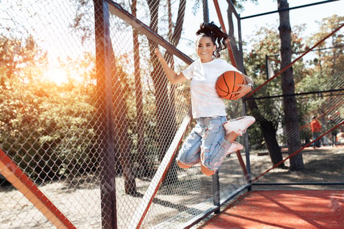 Preview: Careless brunette young woman with bright smile jumping with basketball at the stadium.