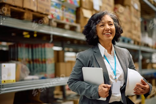 Preview: Happy mature businesswoman in a distribution warehouse looking at camera.
