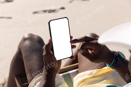 Preview: Midsection of african american senior man using smartphone while relaxing at sandy beach in summer