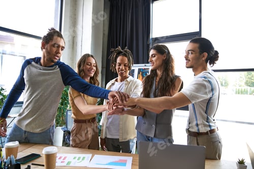 Preview: happy multiethnic business people joining hands near graphs and laptop in office, unity sign
