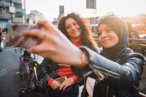 Preview: Young woman in hijab with friend taking smartphone selfie in city