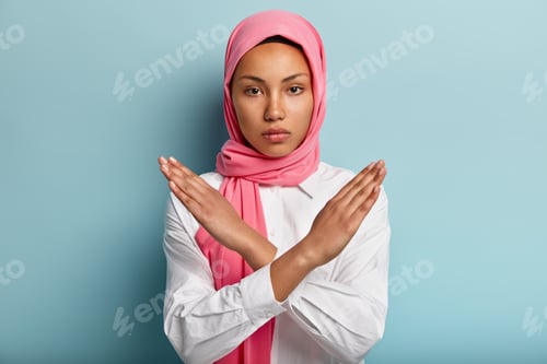Preview: Beautiful serious Arabian woman wears pink hijab, makes cross with hands over chest, shows stop or p