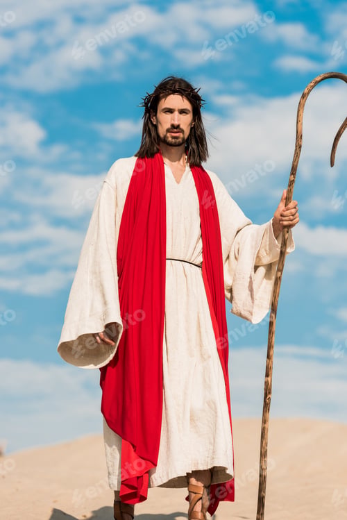 Preview: bearded man in wreath holding wooden cane against blue sky in desert