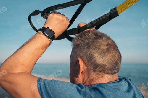 Preview: Older white-haired person performing suspension training on the beach bringing her arms forward