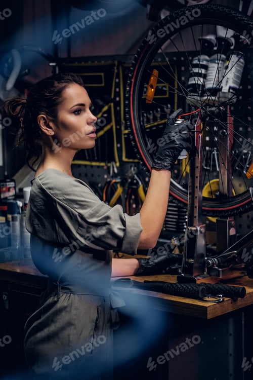Preview: Beautiful woman is repairing bicycle at busy workshop between pneumatic wires.
