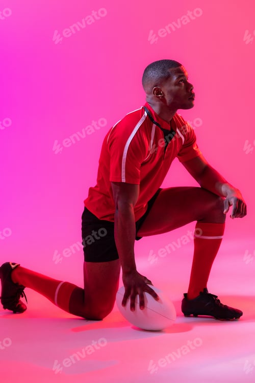 Preview: African american male rugby player crouching with rugby ball over pink lighting