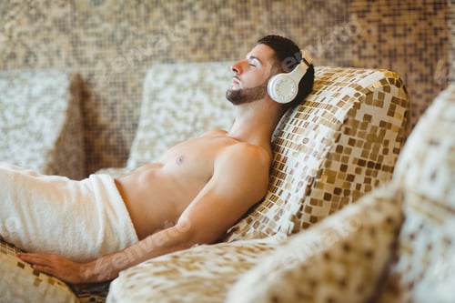 Preview: Male adult reclining on mosaic lounge chair in spa lounge, wearing white towel and headphones