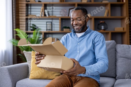 Preview: Smiling man unboxing a package while sitting on a couch at home