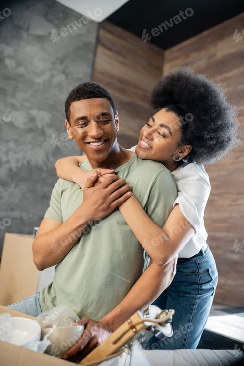 Preview: joyful african american woman hugging boyfriend near cardboard box in new house