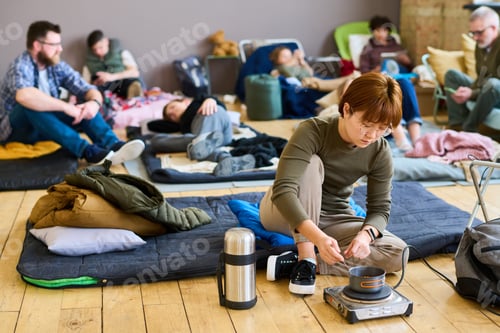 Preview: Young hungry Asian female refugee cooking food in small pan on the floor