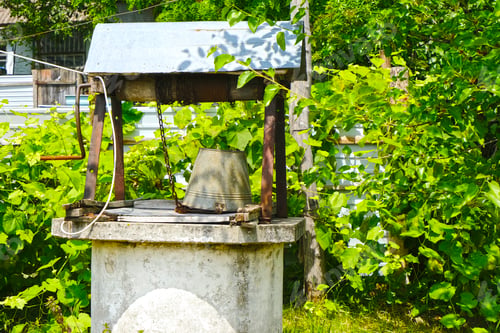 Preview: Old well with a rusty bucket in the yard. Watering. Bright fresh summer greenery view. Countryside.