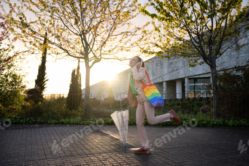 Preview: Businesswoman dancing with rainbow bag and umbrella at sunset