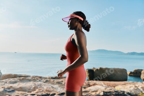 Preview: Woman jogging along the shoreline wearing a red outfit and visor, with a calm sea and distant hills