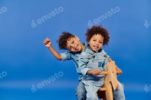 Preview: cute african american siblings in denim clothes sitting together on rocking horse on blue backdrop