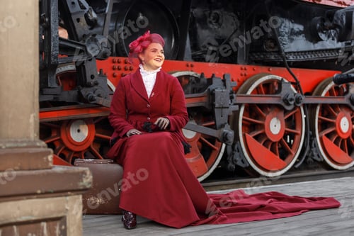 Preview: A beautiful girl in a burgundy suit is sitting on a suitcase near an old steam locomotive