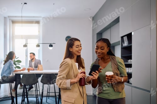 Preview: Happy female entrepreneurs using cell phone on coffee break during work hours in office kitchen.