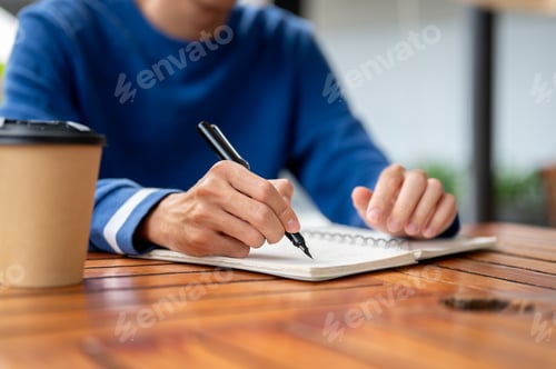 Preview: Close up of a hand holding pen writing in notebook aside coffee at wooden table in cafe or canteen.