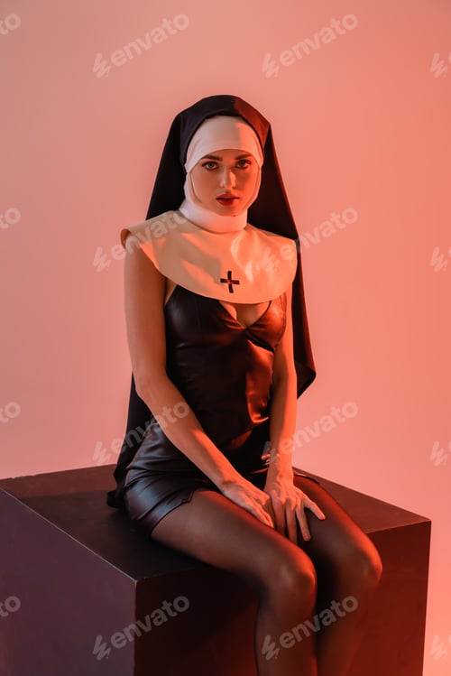 Preview: sexy nun in leather dress looking at camera while sitting on black cube isolated on pink