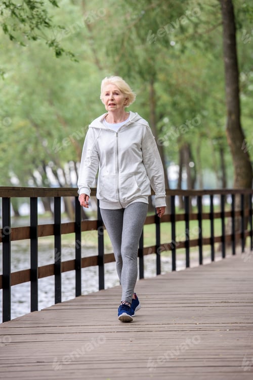 Preview: senior woman in sportswear walking on wooden path in park near river