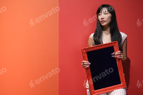 Preview: Young asian woman pose with a red framed mirror over a orange background.