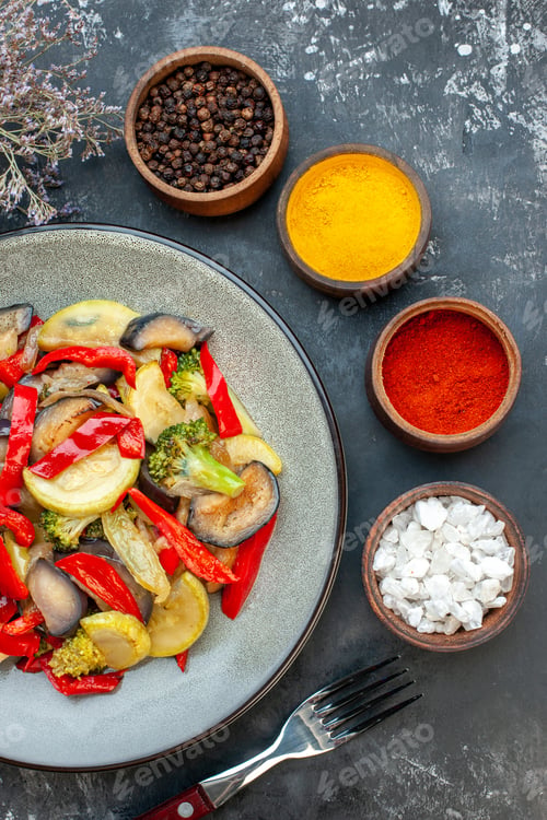 Preview: Vertical view of delicious vegetarian meal on a plate and fork next to different spices on dark