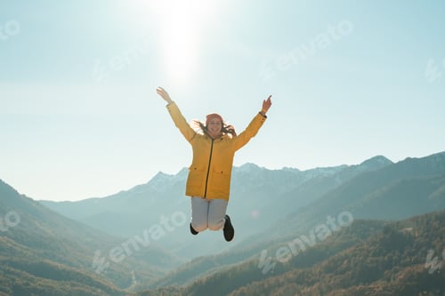 Preview: Young woman in hat and yellow jacket jumps for happiness in mountains on sunny autumn day.