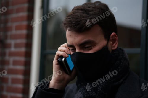 Preview: Handsome young man making a phone call, smartphone at his ear, wearing a face mask and smiling.