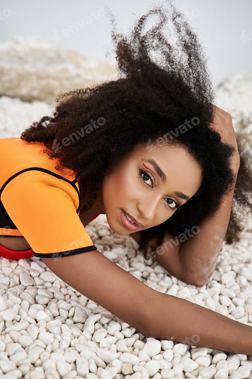 Preview: Stylish young African American woman poses gracefully among smooth pebbles in a vibrant outfit