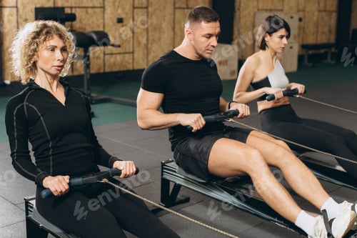 Preview: Young man and beautiful women working out with rowing machine at crossfit gym.
