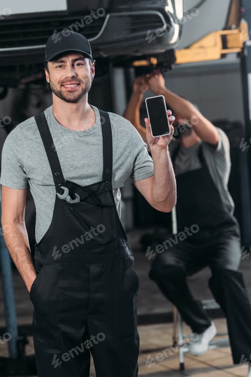 Preview: auto mechanic showing smartphone with blank screen, while colleague working in workshop behind