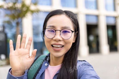 Preview: A smiling asian woman with glasses waves hello, looking directly at the camera outside.