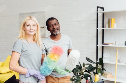 Preview: smiling blonde woman and african american man in rubber gloves holding dusters