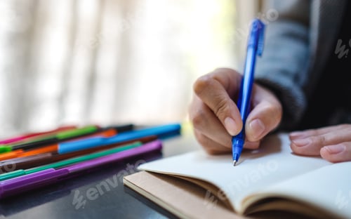 Preview: Closeup image of a woman writing on a blank notebook with colored pens on the table
