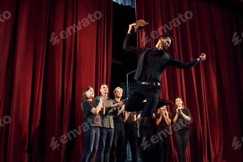 Preview: Standing against red curtains. Group of actors in dark colored clothes on rehearsal in the theater