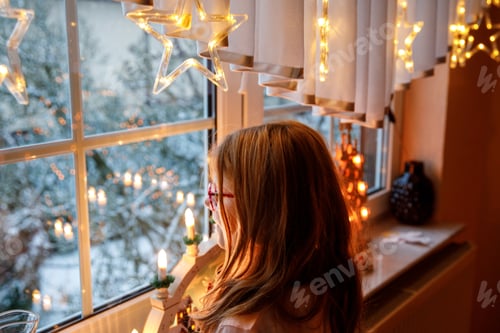 Preview: little girl sitting by the window and looking at the winter snowing landscape.