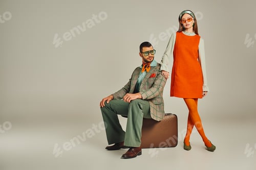 Visualização: homem moderno sentado em uma mala vintage perto de mulher em vestido laranja em casal cinza de inspiração retrô