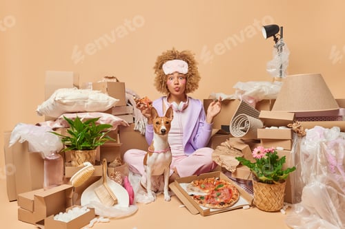 Preview: Moving day concept. Impressed curly woman relocates in new apartment together with her pet eats pizz